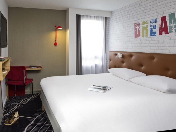 Une chambre d’hôtel avec un grand lit, un fauteuil rouge, un bureau avec une lampe, une télévision et un mur en briques blanches sur lequel est écrit le mot « CREAM ».