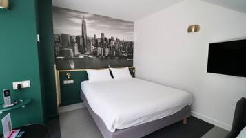 A hotel room with a bed, a desk with a phone, a TV, and a cityscape mural on the wall.