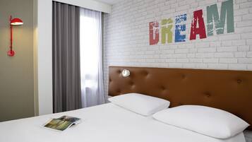 A hotel room with a white brick wall, a large bed with two white pillows, a red floor lamp, and a window with grey curtains.