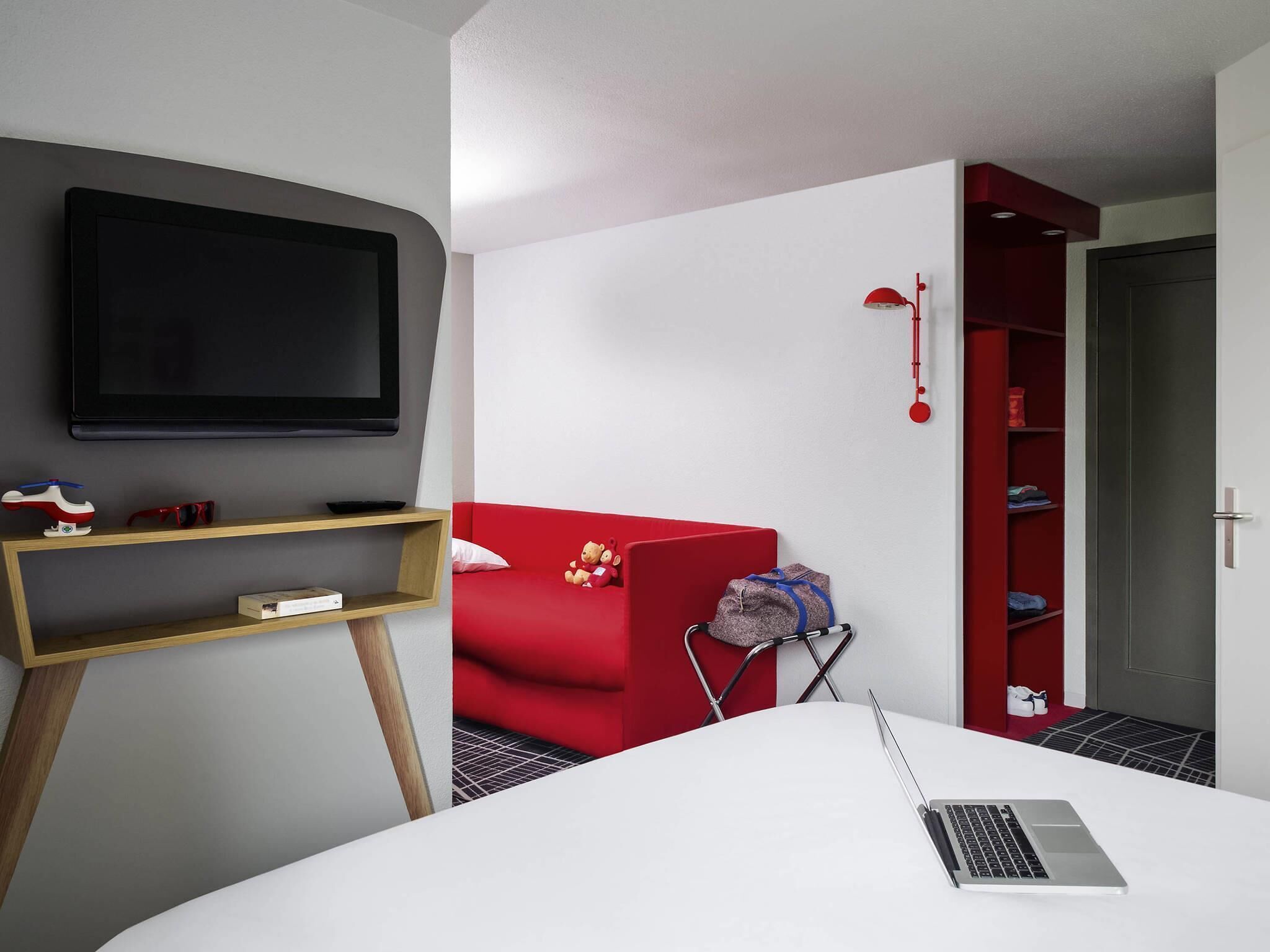 Ein modernes Zimmer mit Fernseher, einem roten Sofa, einem weißen Schreibtisch mit Laptop und einem roten Kleiderschrank.