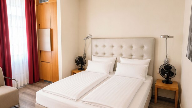 Una cama doble con sábanas blancas, dos mesitas de noche con ventiladores, un armario de madera y una cortina roja.