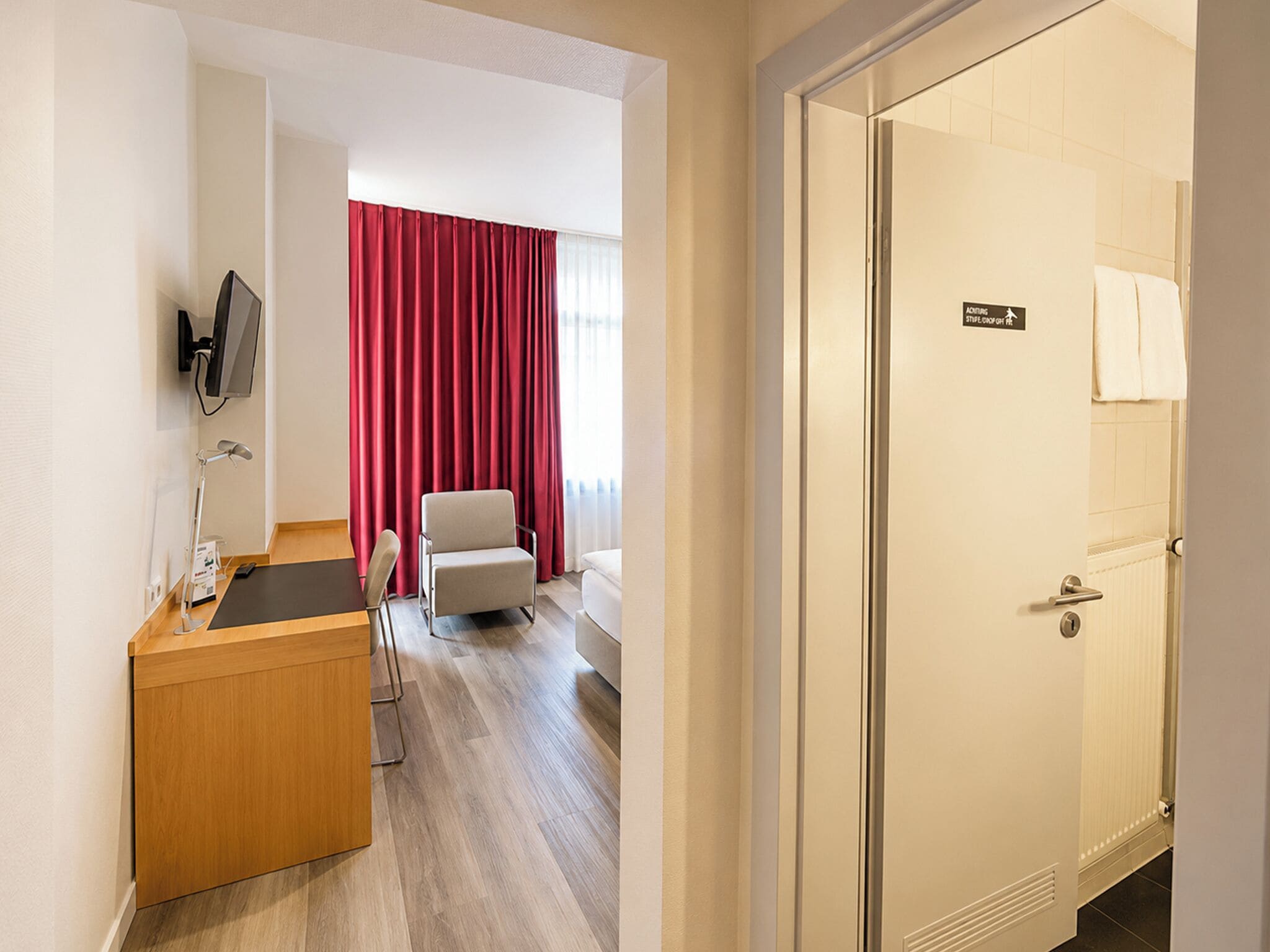 A hotel room with a wooden desk, a chair, a bed, a TV mounted on the wall, and a door with a sign.