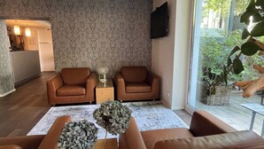 Una sala de estar con dos sofás de cuero marrón, una mesa de centro con flores decorativas, un televisor montado en la pared y vistas a zonas verdes al exterior.