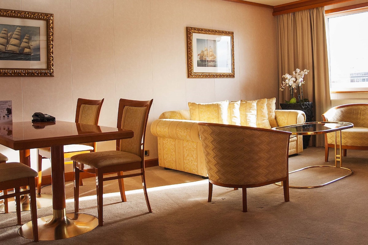 Quarto de hotel com área de refeições, sofá e janela com cortinas.