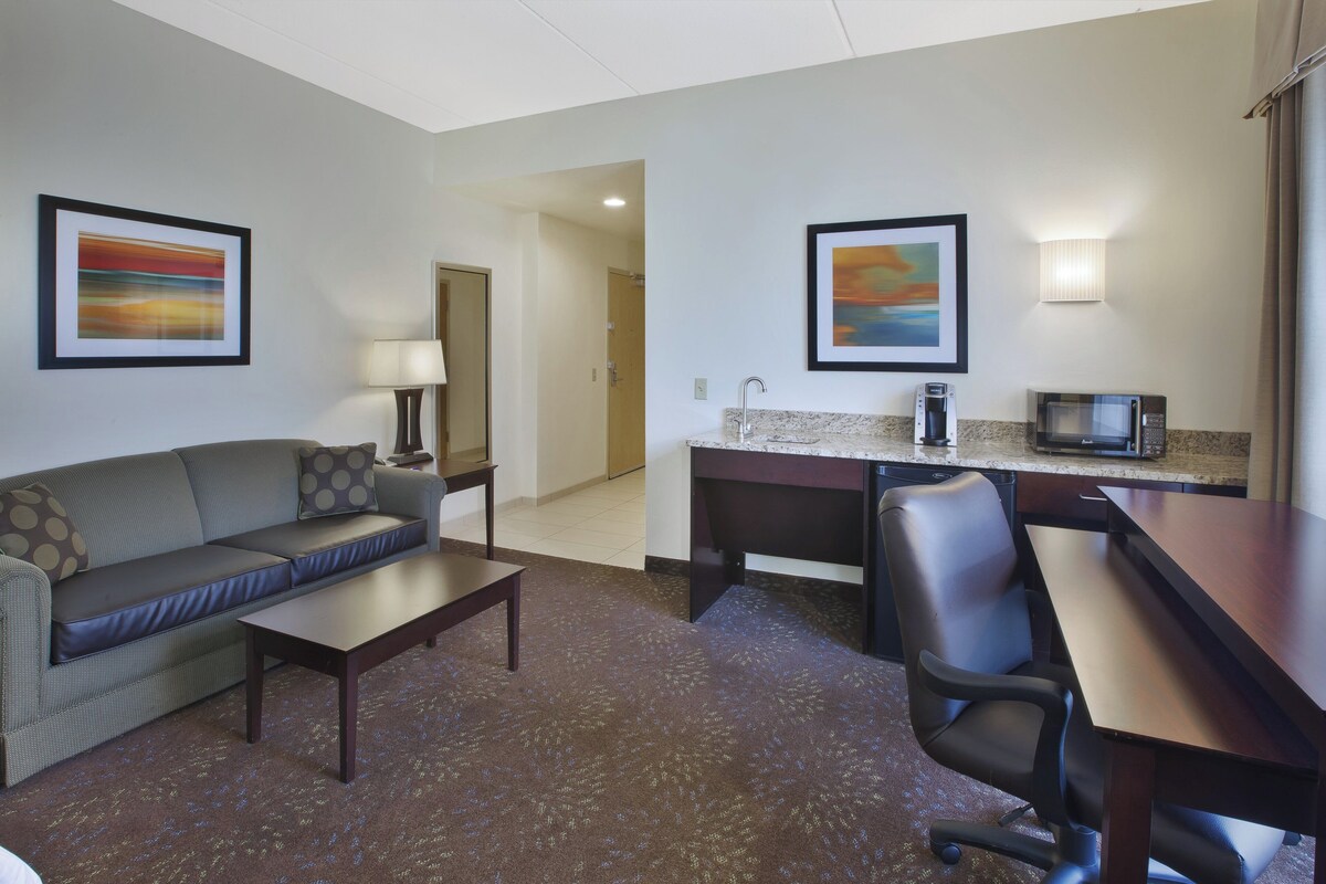 A hotel room with a sofa, a small table, a desk with a chair, a microwave, and a coffee maker. There are two framed pictures on the walls.