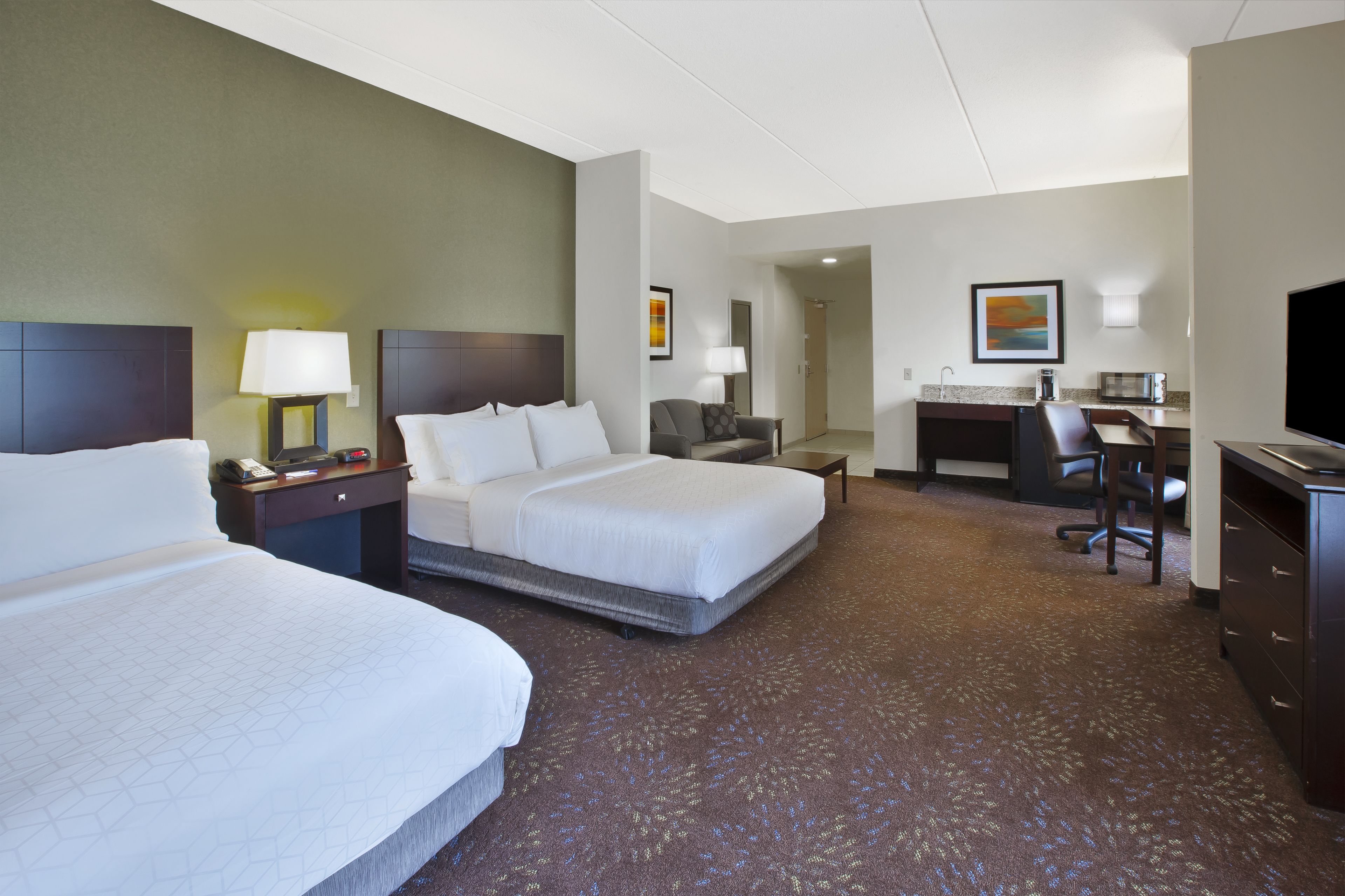 A hotel room with two beds, a desk, a chair, a TV, and a kitchenette.