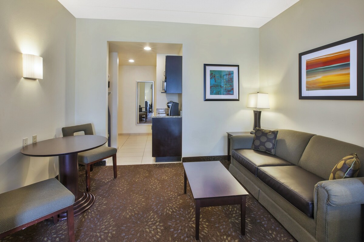 A hotel room with a dining table, a sofa, a coffee table, a lamp, and artwork on the walls.