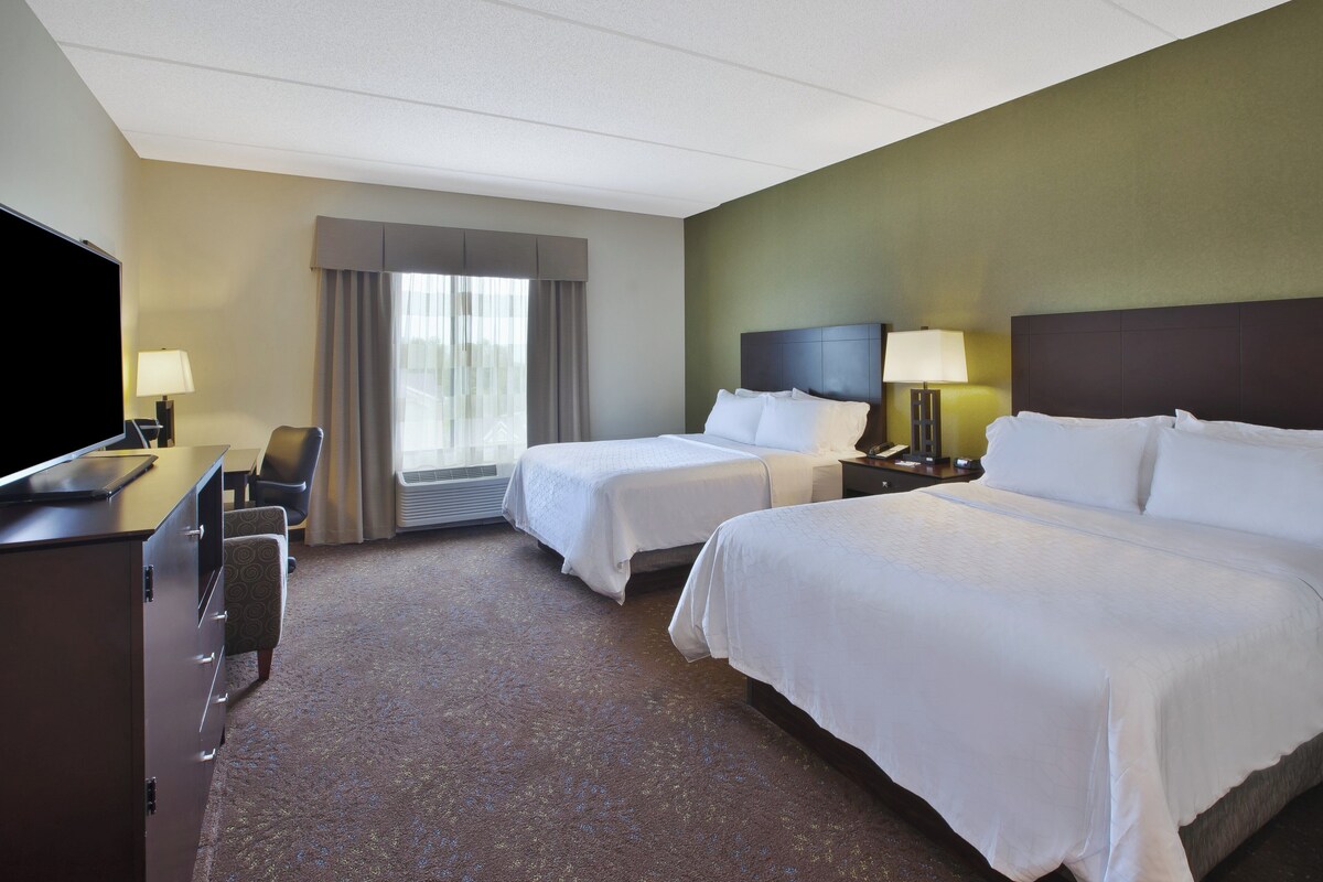 A hotel room with two beds, a desk with a TV, a chair, a nightstand with a lamp, and a window with curtains.