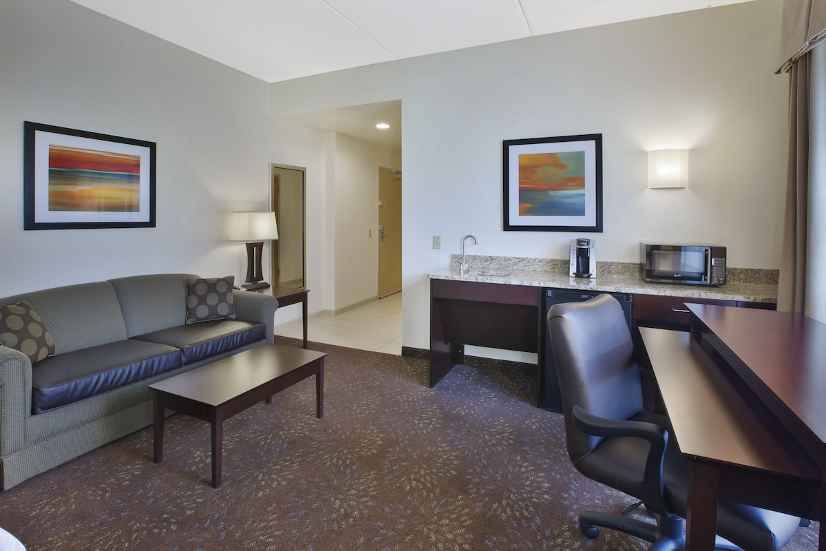 A hotel room with a sofa, coffee table, desk, and microwave. There are framed pictures on the walls and a lamp on the desk.