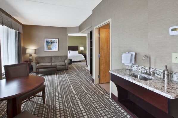 A hotel room with a dining table, a sofa, a bed, a bathroom with a sink and mirror, and a patterned floor. - Holiday Inn Express & Suites Geneva Finger Lakes by IHG (Geneva)