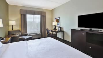 Habitación de hotel con una cama grande, un escritorio, un televisor de pantalla plana, una silla, una lámpara y una ventana con cortinas.