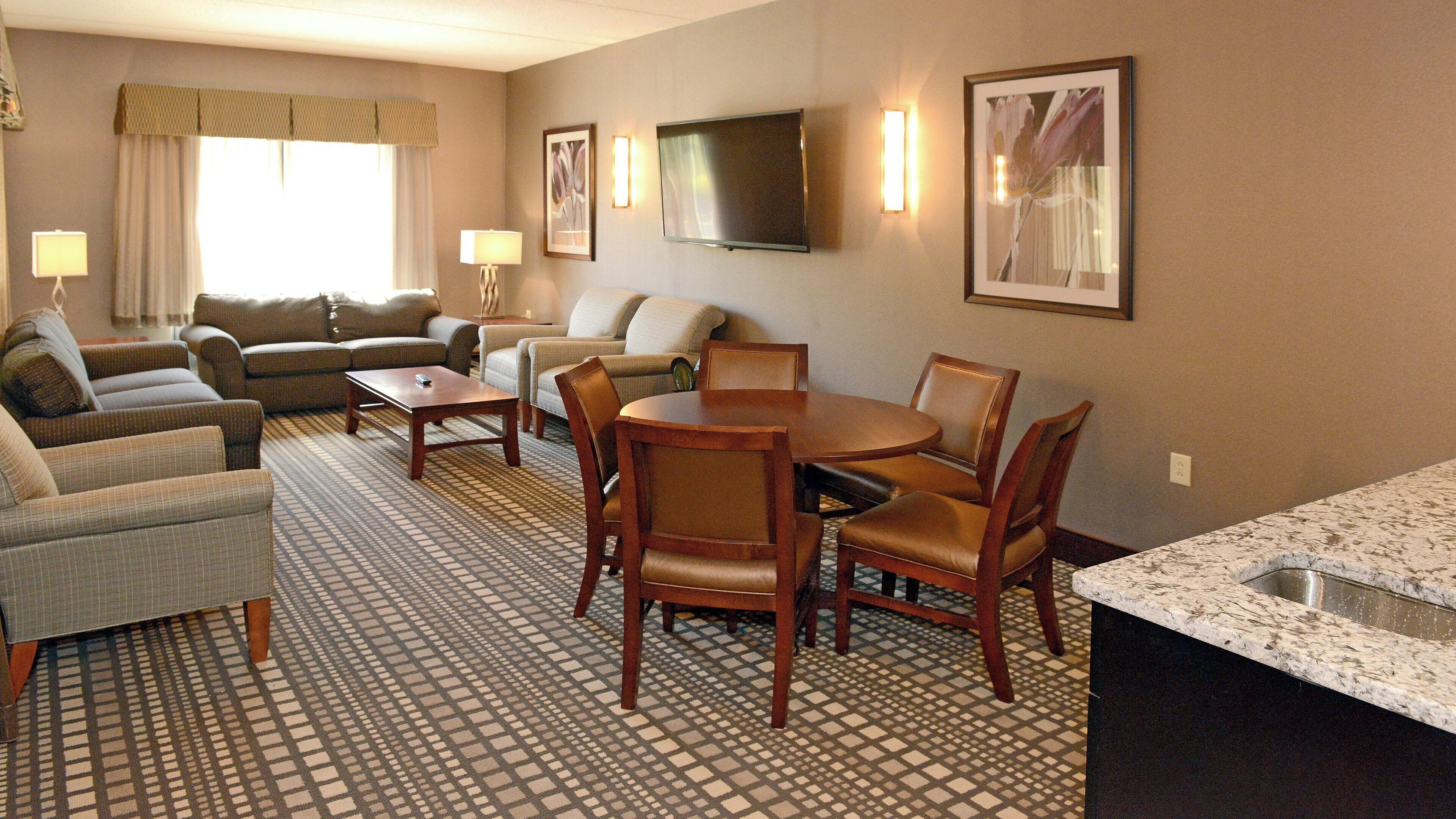 A hotel room with a living area, a dining table with chairs, a flat-screen TV mounted on the wall, and a patterned carpet.