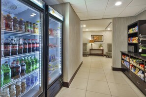 A hotel corridor with a refrigerator stocked with beverages, a seating area with a lamp, and snack shelves.