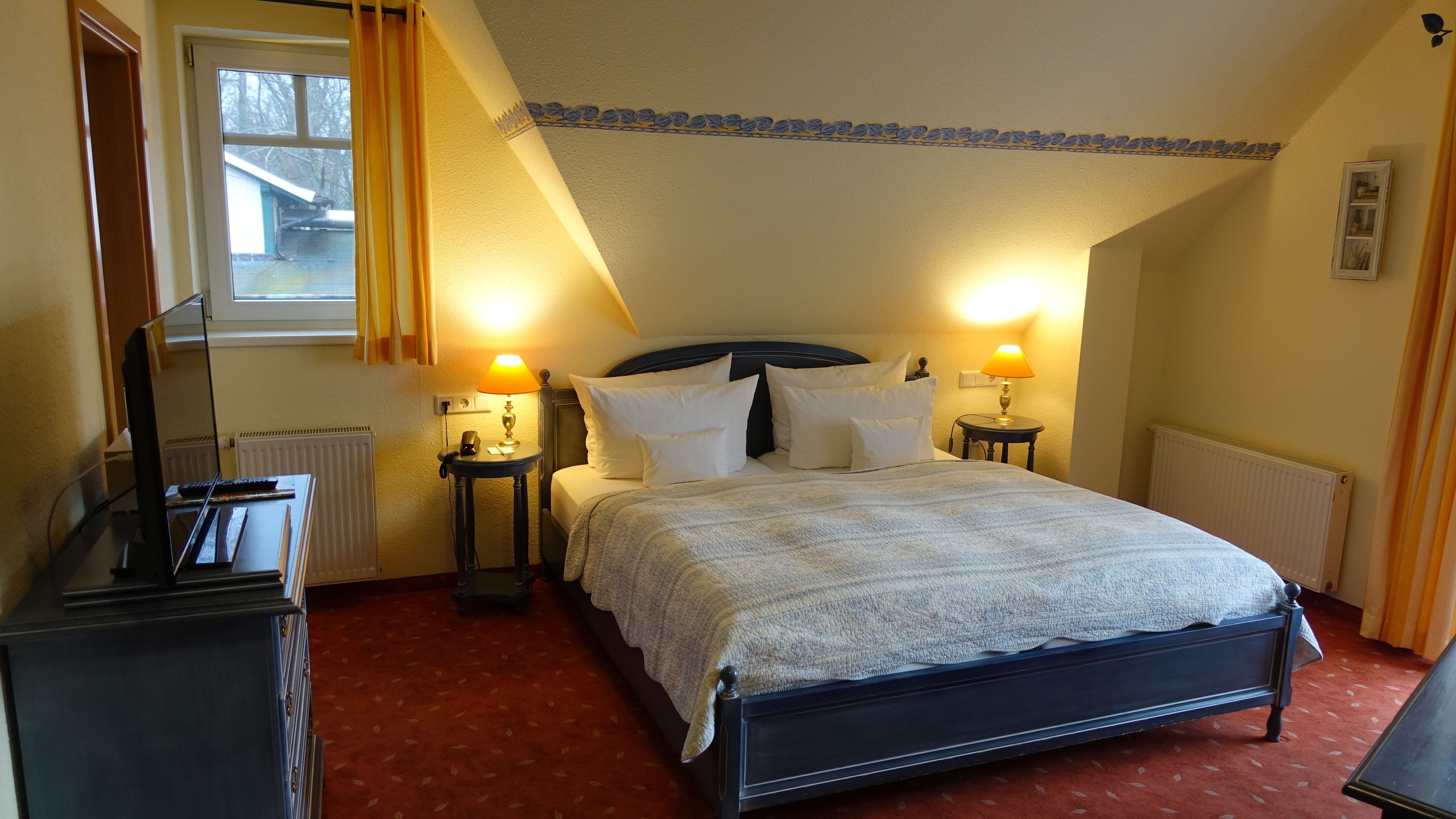 A hotel room with a bed, two bedside tables with lamps, a dresser, a radiator, and a window with curtains.