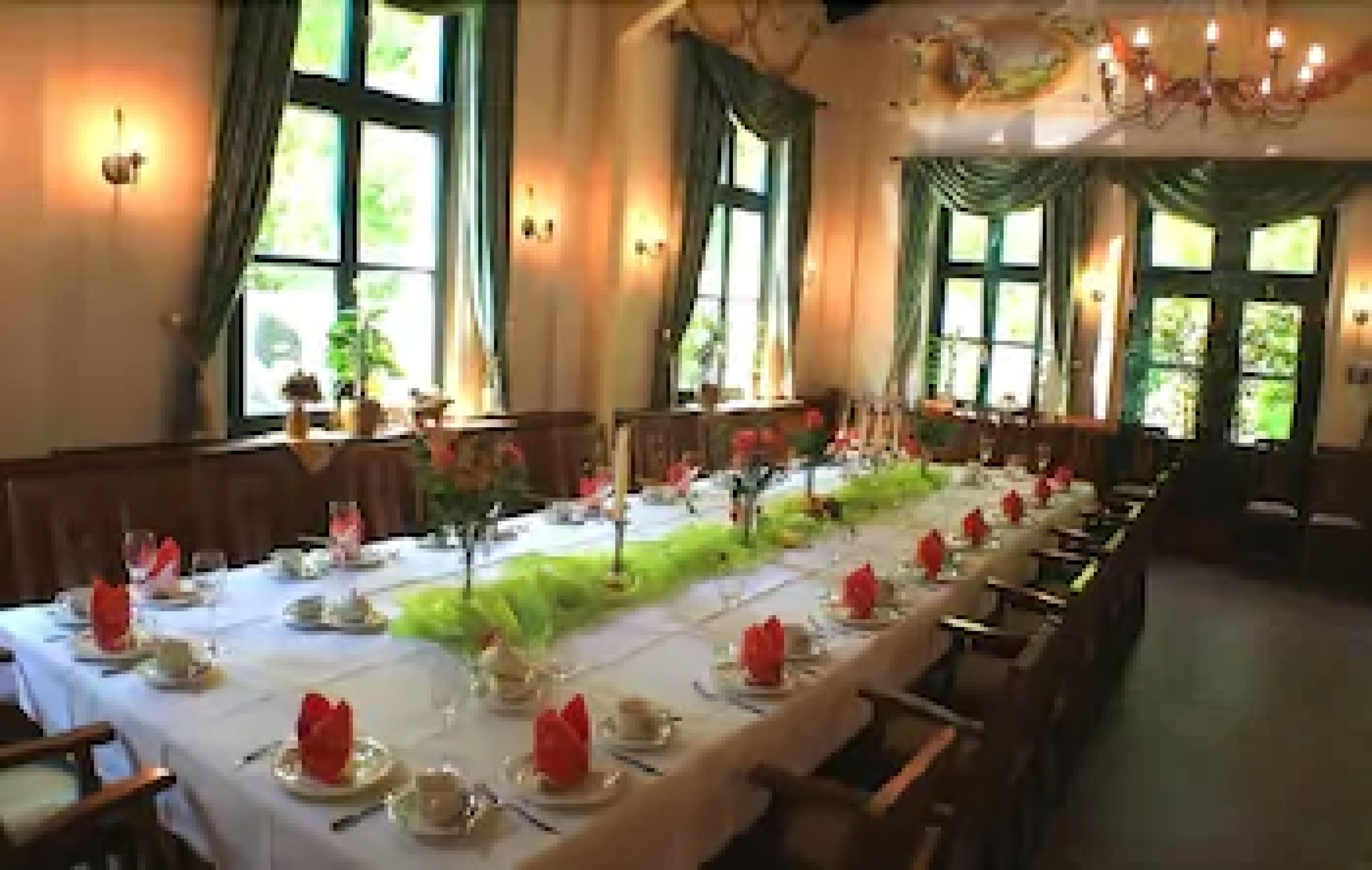 A long dining table set with red napkins, plates, and glasses, flanked by windows with curtains.