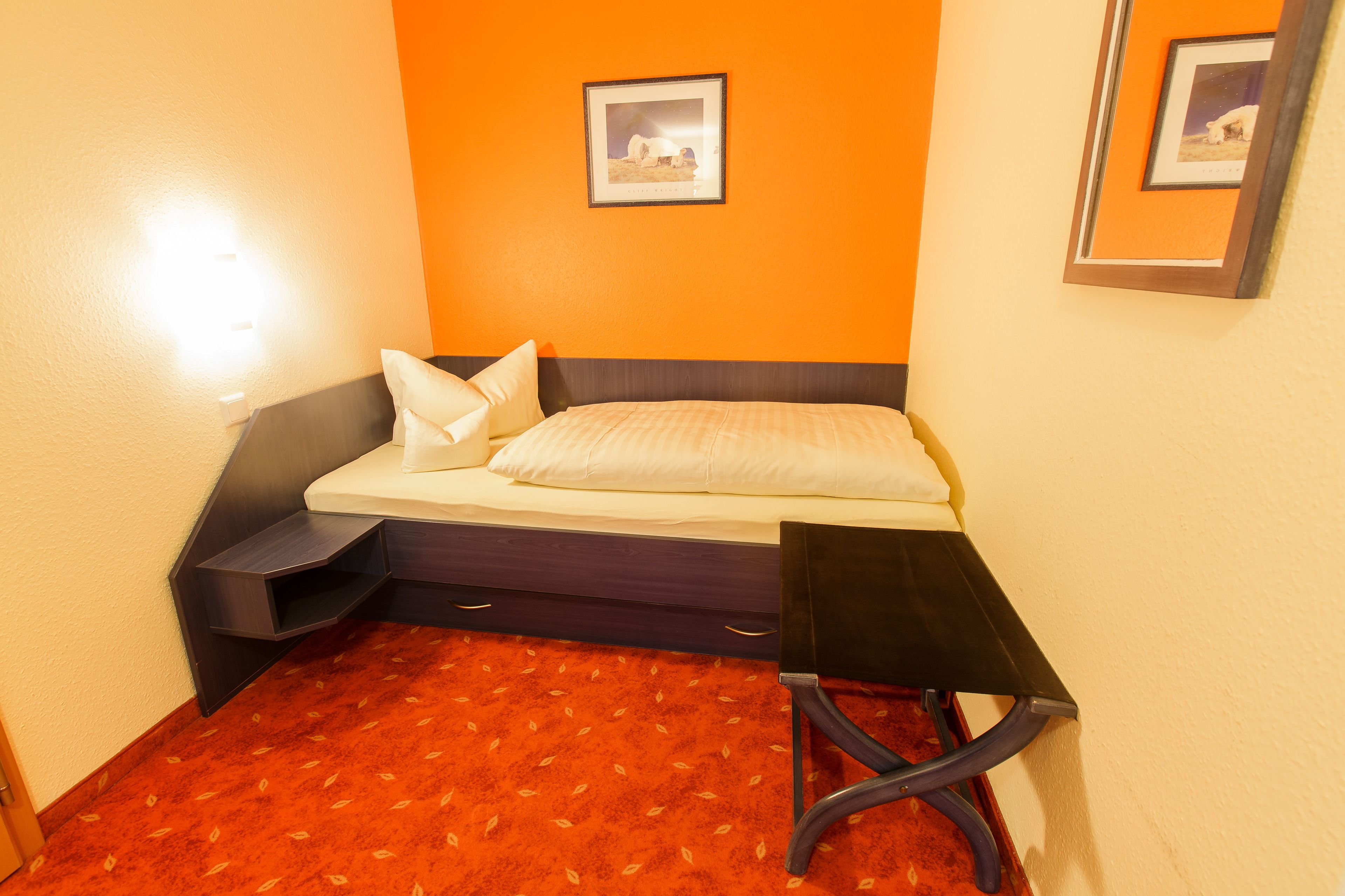 A compact hotel room with a bed, a small bedside table, a chair, and a carpeted floor.