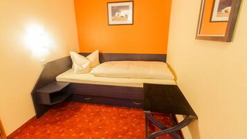 A compact hotel room with a bed, a small bedside table, a chair, and a carpeted floor.