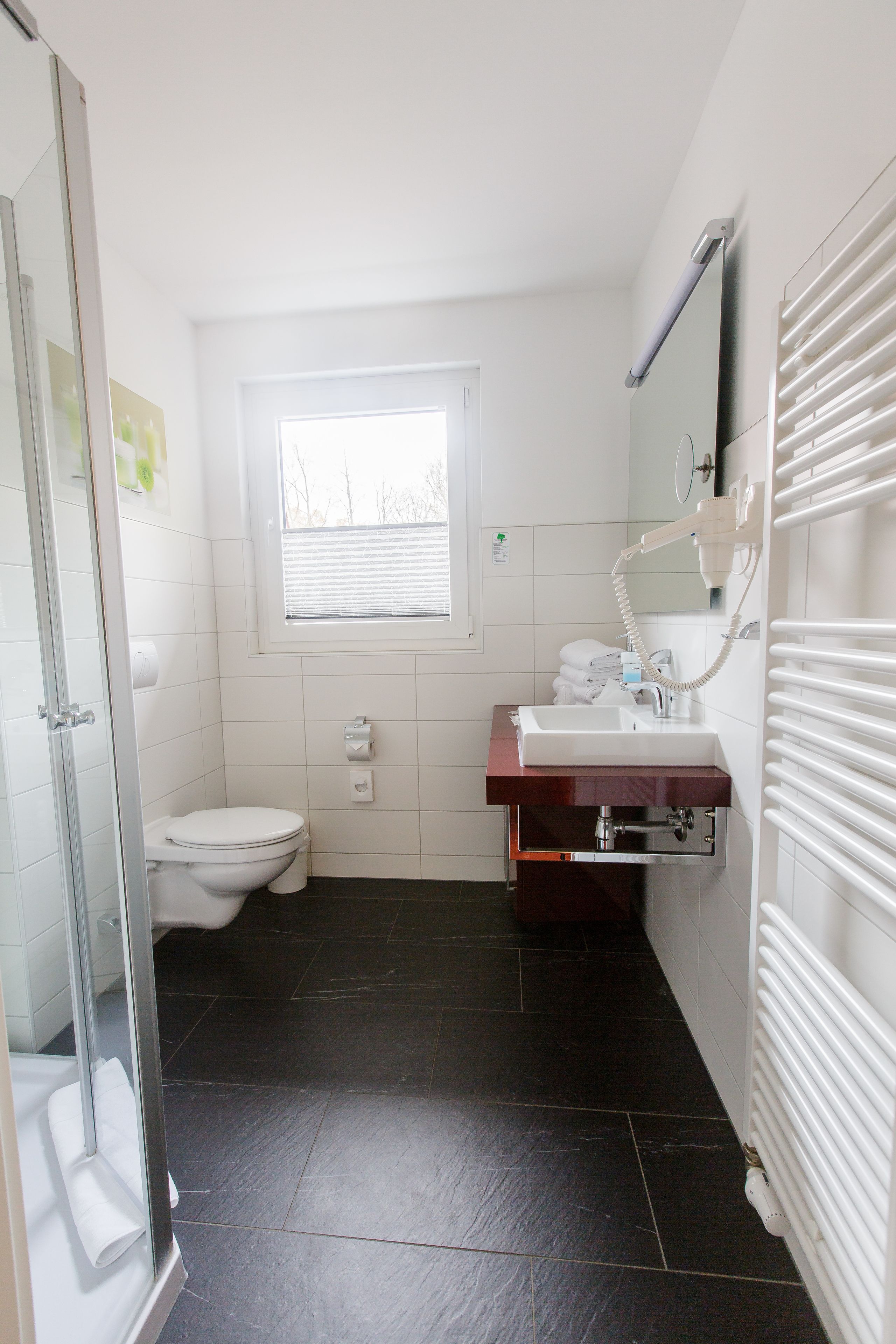 Ein Badezimmer mit Duschabtrennung aus Glas, einer weißen Toilette, einem Waschbecken mit Spiegel und einem Fenster mit Jalousien.