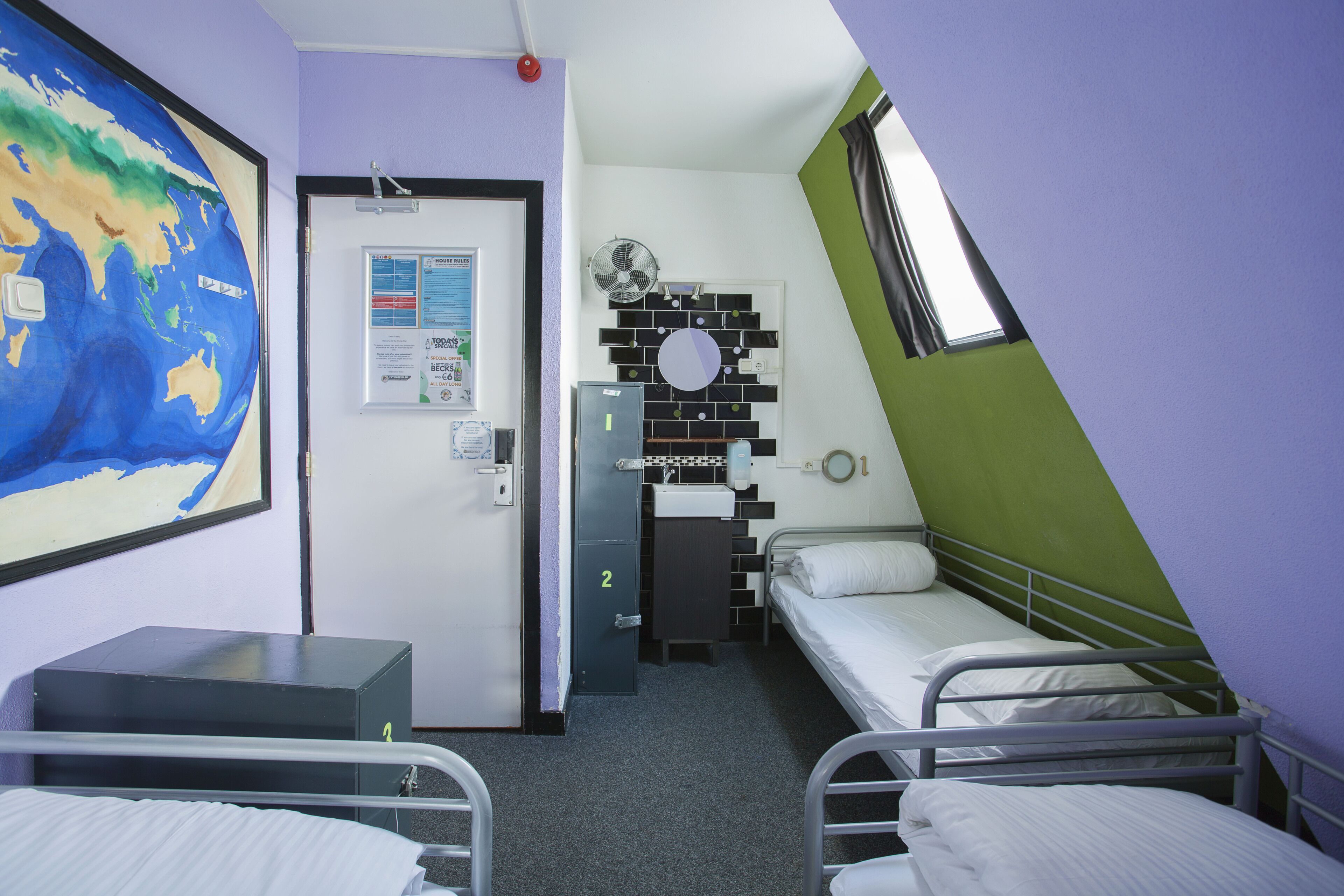 Een kleine, compacte kamer met twee eenpersoonsbedden, een bureau, een stoel, een wereldkaart aan de muur, een deur met een nummer en een klein raam.