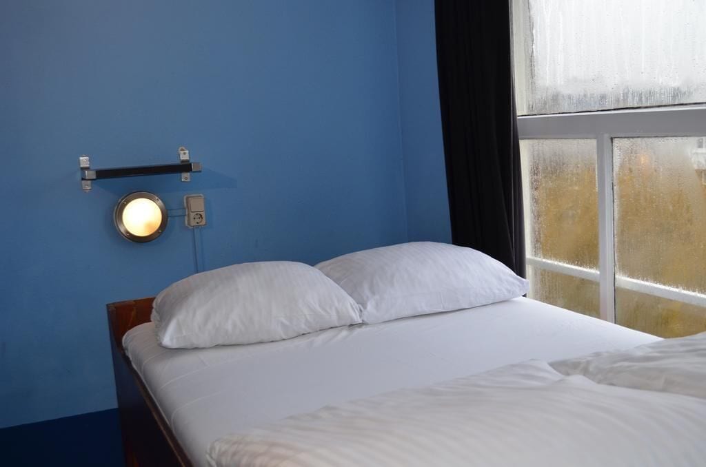 Een slaapkamer met een bed, witte kussens, een wandlamp en een raam met gordijnen.