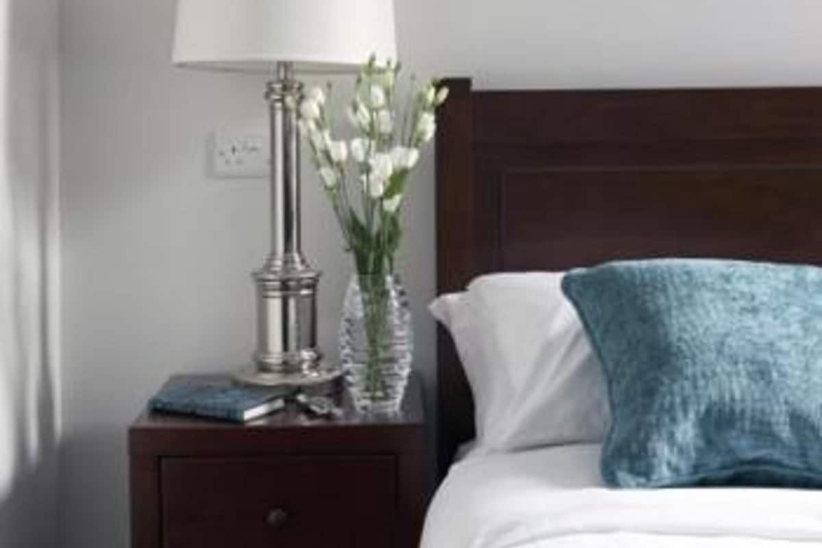 A neatly made bed with a white comforter and a blue throw pillow, a bedside table with a lamp and a vase of flowers, and a framed picture on the wall.