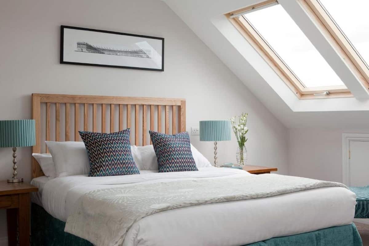 A bedroom with a sloped ceiling, a bed with white bedding and patterned pillows, two bedside tables with lamps, and a framed picture on the wall.
