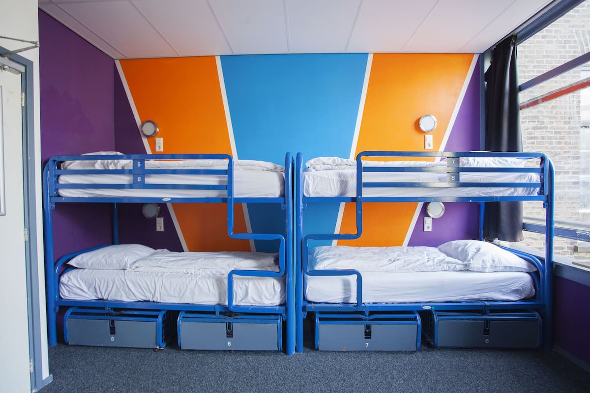 Een slaapzaal met twee stapelbedden, elk voorzien van een matras en kussens, en een klein opbergkastje onder elk bed.
