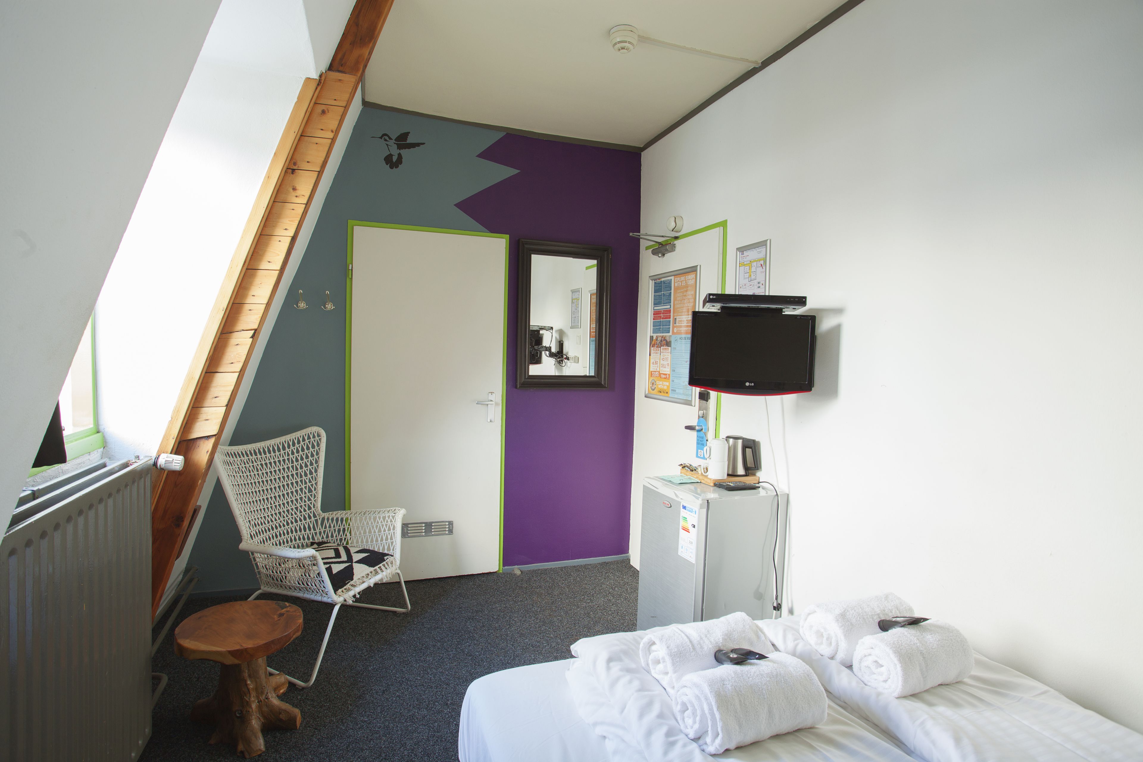 A hotel room with a bed, a small wooden stool, a white chair, a desk with a computer and a kettle, and a purple accent wall.