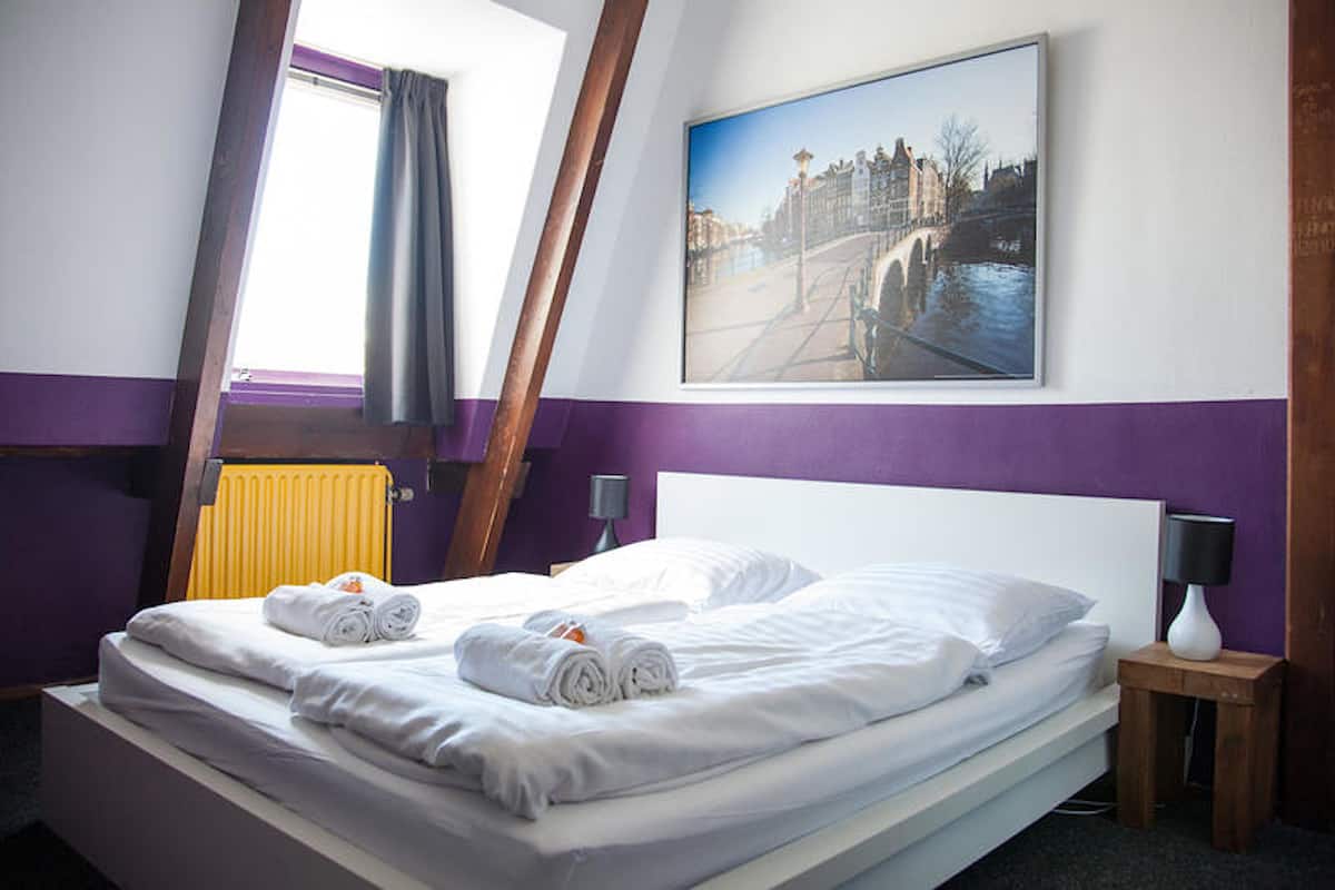 Een netjes opgemaakt bed met opgerolde handdoeken, een schilderij van een brug, een raam met gordijnen en een bedlampje.
