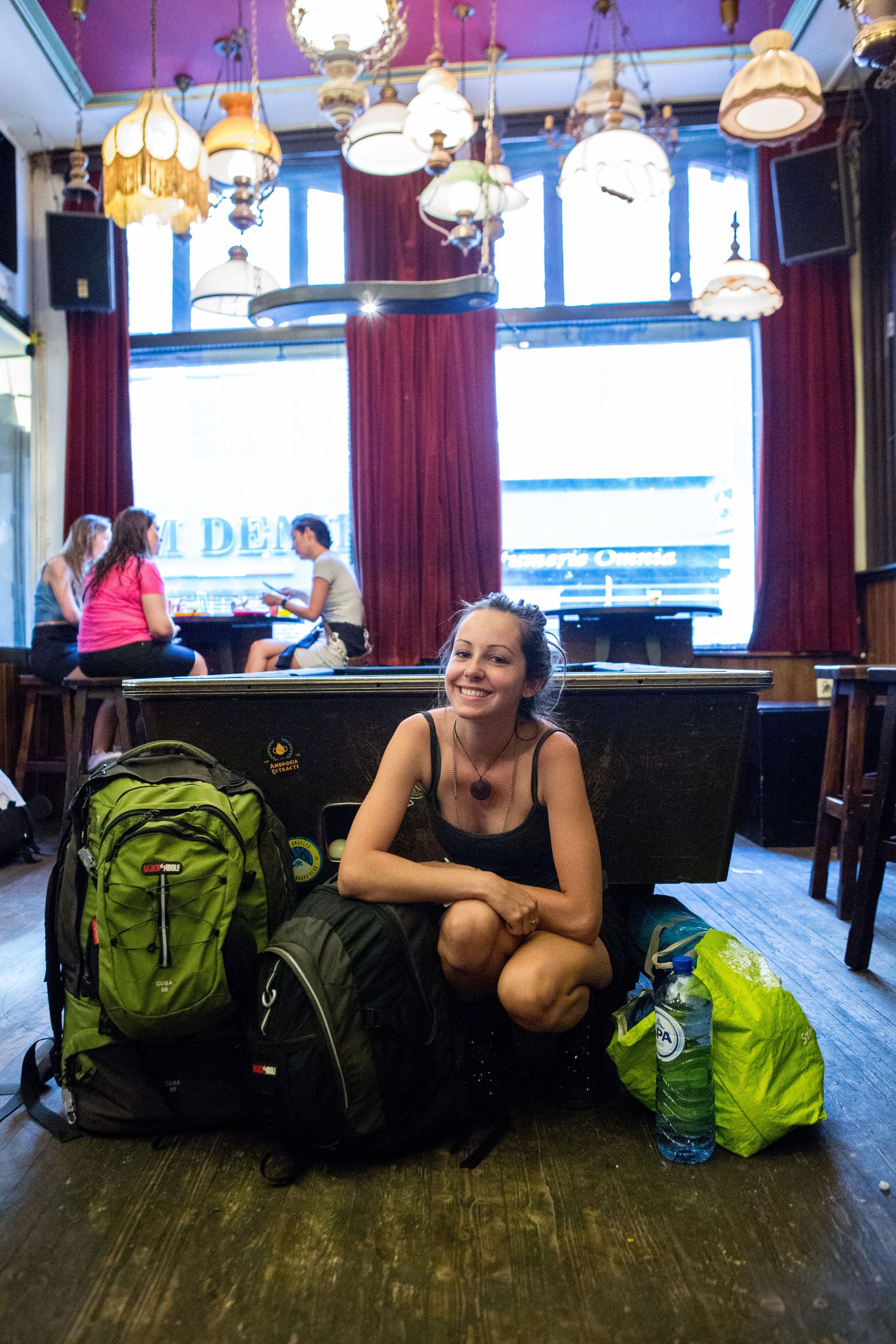 A woman sitting on a bench with a green backpack and a water bottle beside her in a room with wooden flooring and hanging lamps.