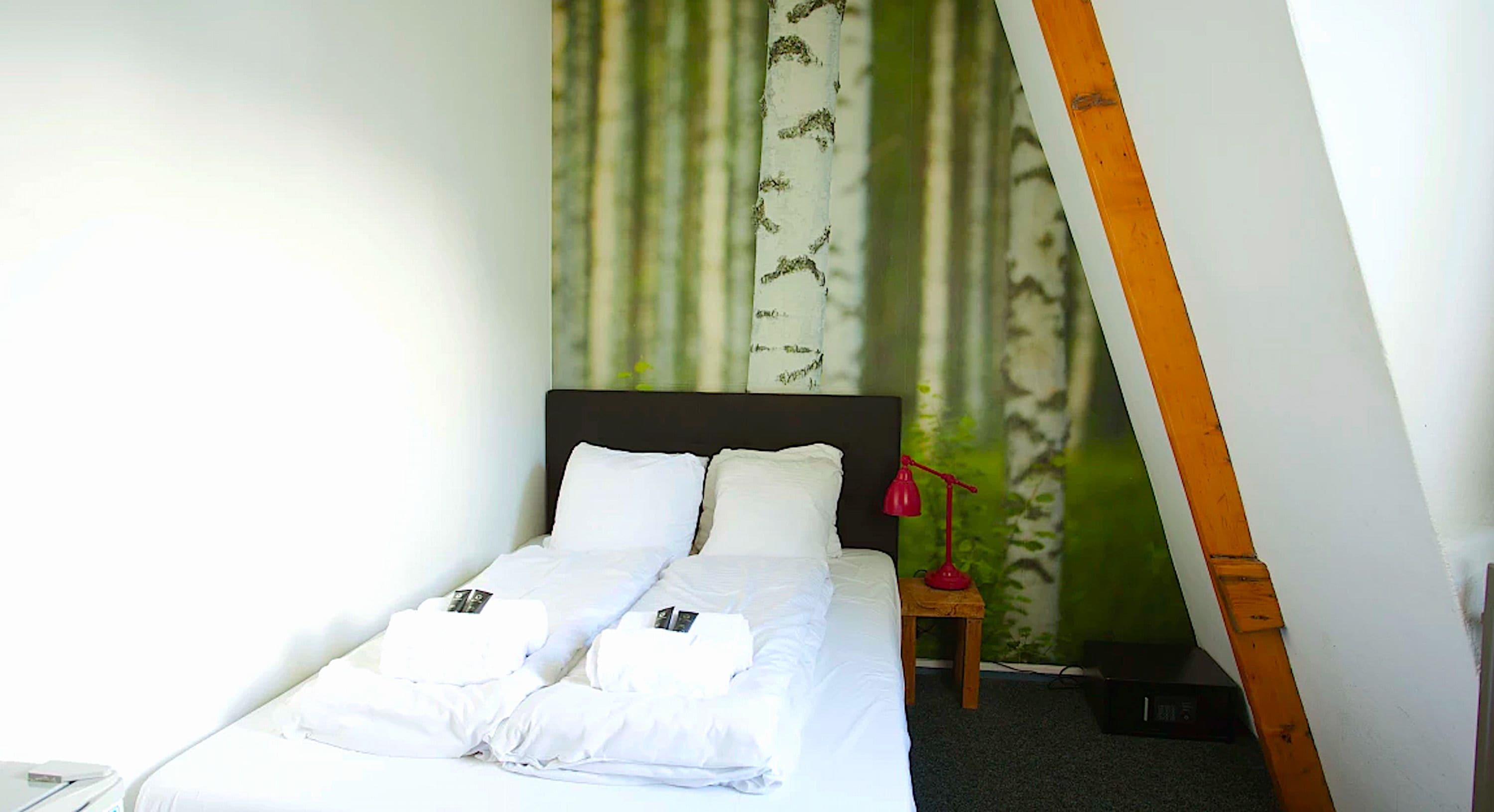 Una cama bien hecha con ropa de cama y toallas blancas, una lámpara de escritorio roja y una cortina de pared con estampado verde y blanco.