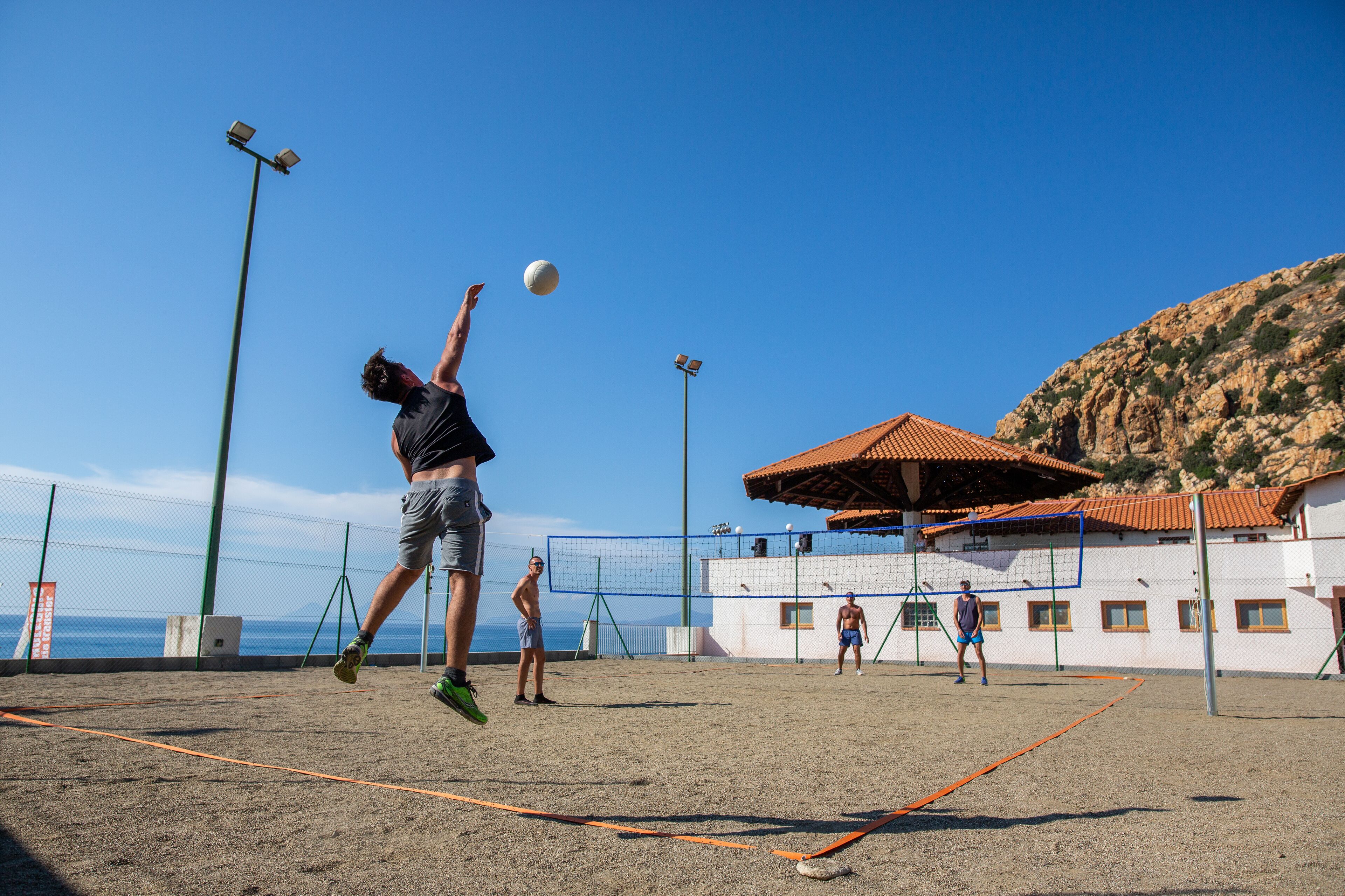 A beach volleyball court with a net, players, and a building with a red-tiled roof in the background.