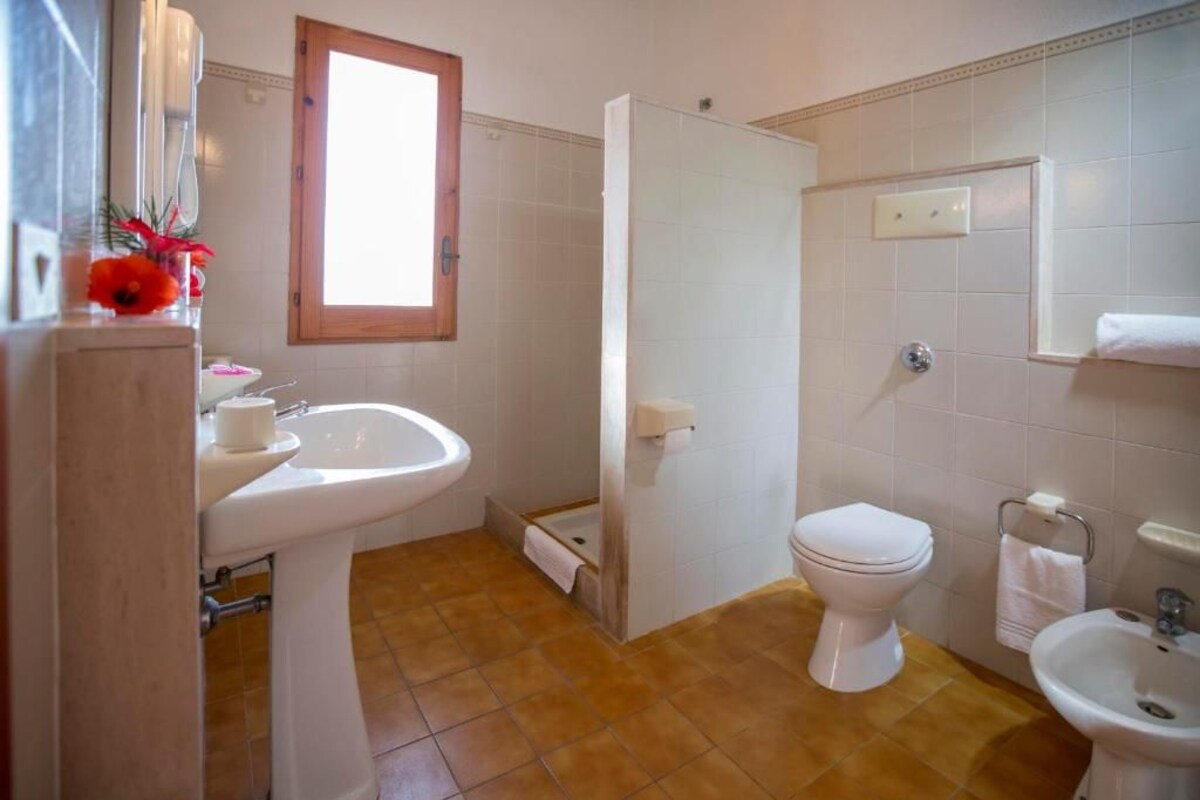 Un baño con lavamanos, inodoro y bidé blancos, una ventana con marco de madera y piso de baldosas.
