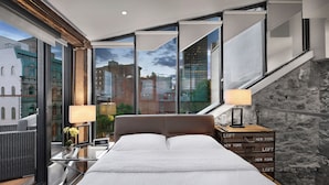 Un dormitorio moderno con una cama grande, una mesita de noche y un aparador. Amplias ventanas ofrecen vistas de la ciudad.