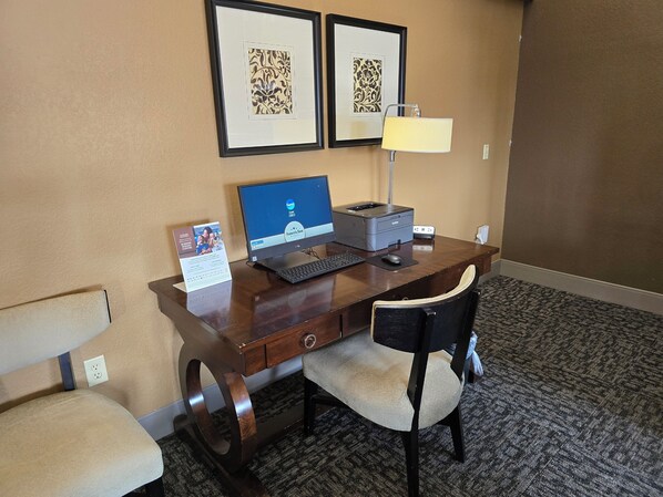 A hotel room with a wooden desk, a laptop, a printer, a lamp, and two framed pictures on the wall.