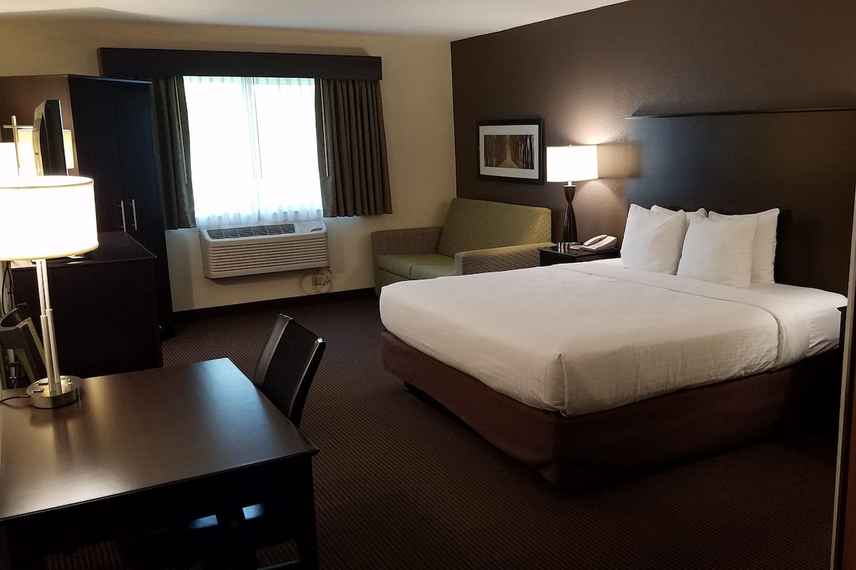 A hotel room with a large bed, a desk, a chair, a lamp, a phone, a picture on the wall, and an air conditioning unit.