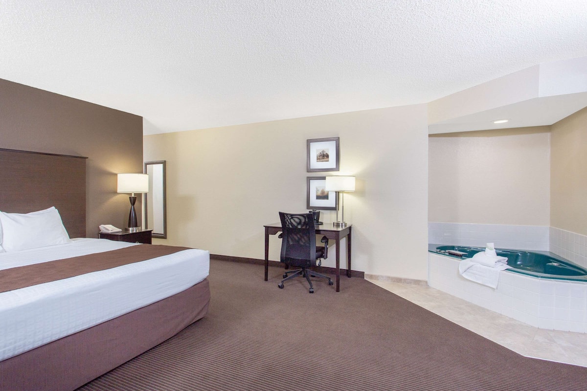 A hotel room with a large bed, a desk with a chair, a desk lamp, a telephone, and a bathtub.