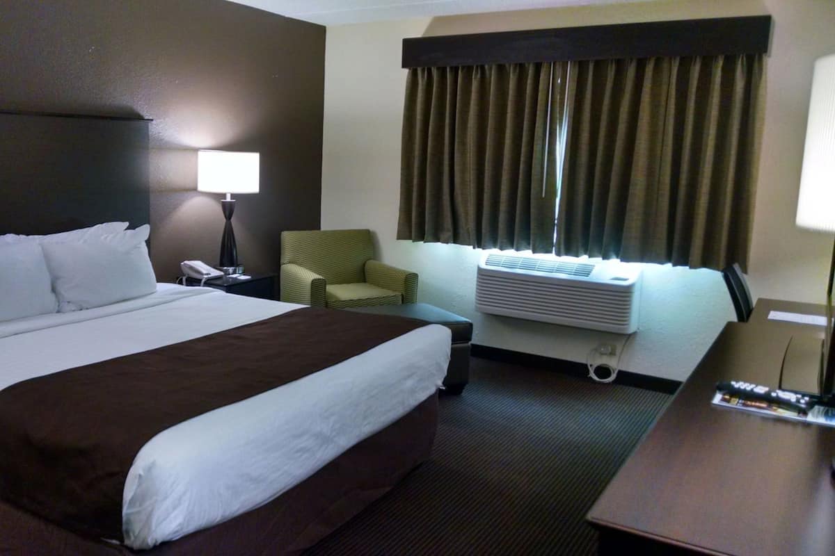 A hotel room with a bed, a chair, a desk, a lamp, an air conditioner, and a window with curtains.