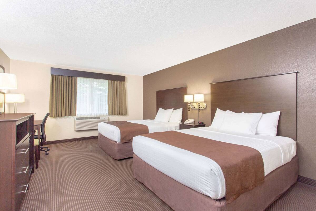 A hotel room with two beds, a desk, a chair, a lamp, and a window with curtains.