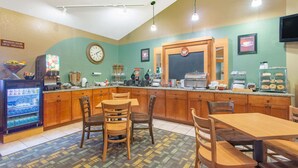 A breakfast area with wooden tables and chairs, a refrigerator with beverages, a clock, and a coffee machine.