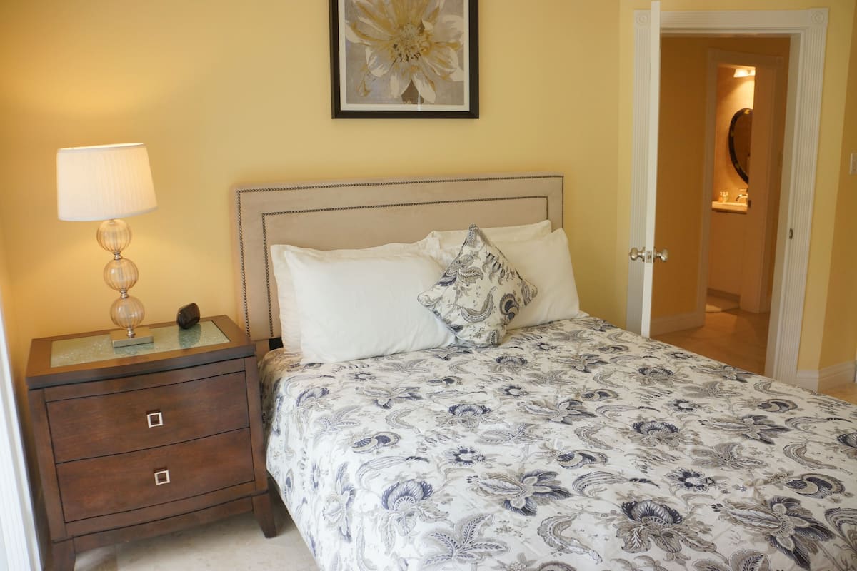 Een net opgemaakt bed met een bloemenprint sprei, een bedlampje en een lijst met bloemenkunst aan de muur.