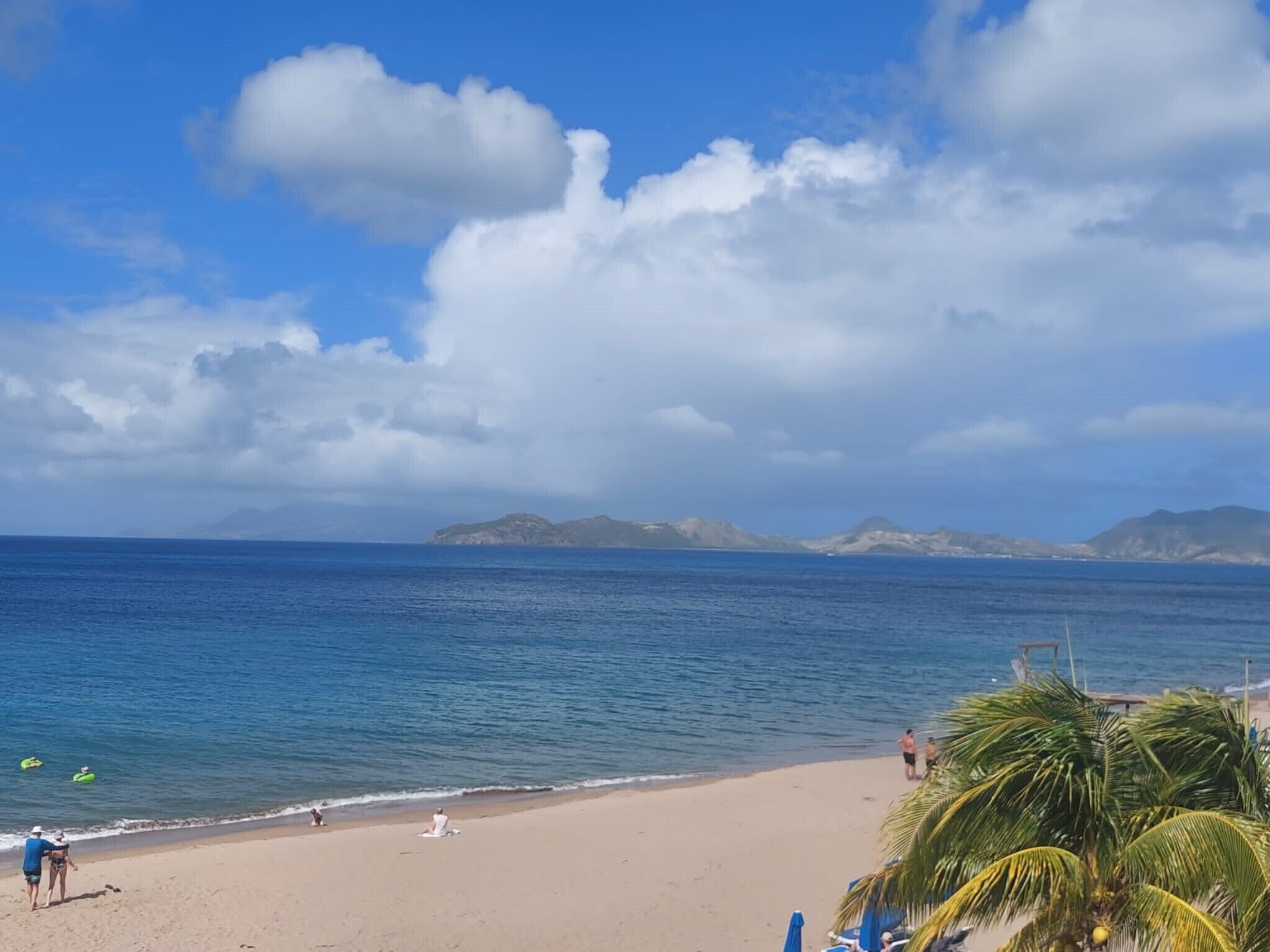 A beach with clear skies, calm sea, and distant mountains.