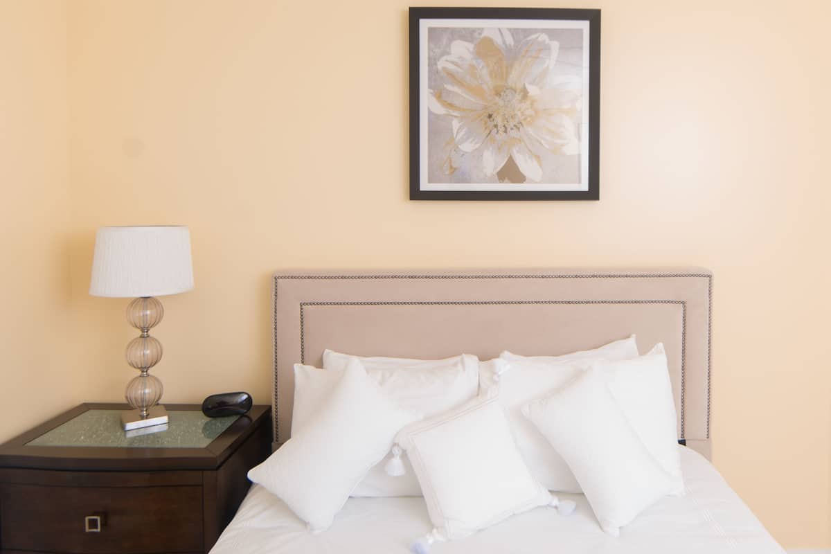 Een net opgemaakt bed met witte beddengoed en kussens, een bedlampje, een lijst met bloemkunst aan de muur, en een donker houten nachtkastje met een boek en een telefoon.