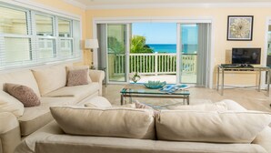 A living room with a beige sofa, a glass coffee table, and a TV on a stand. There is a balcony with a view of the ocean and a white fence. - The Hamilton Beach Villas & Spa (Cotton Ground)