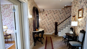 Un pasillo con suelo alfombrado, una escalera de madera, una mesa con lámpara y una silla.
