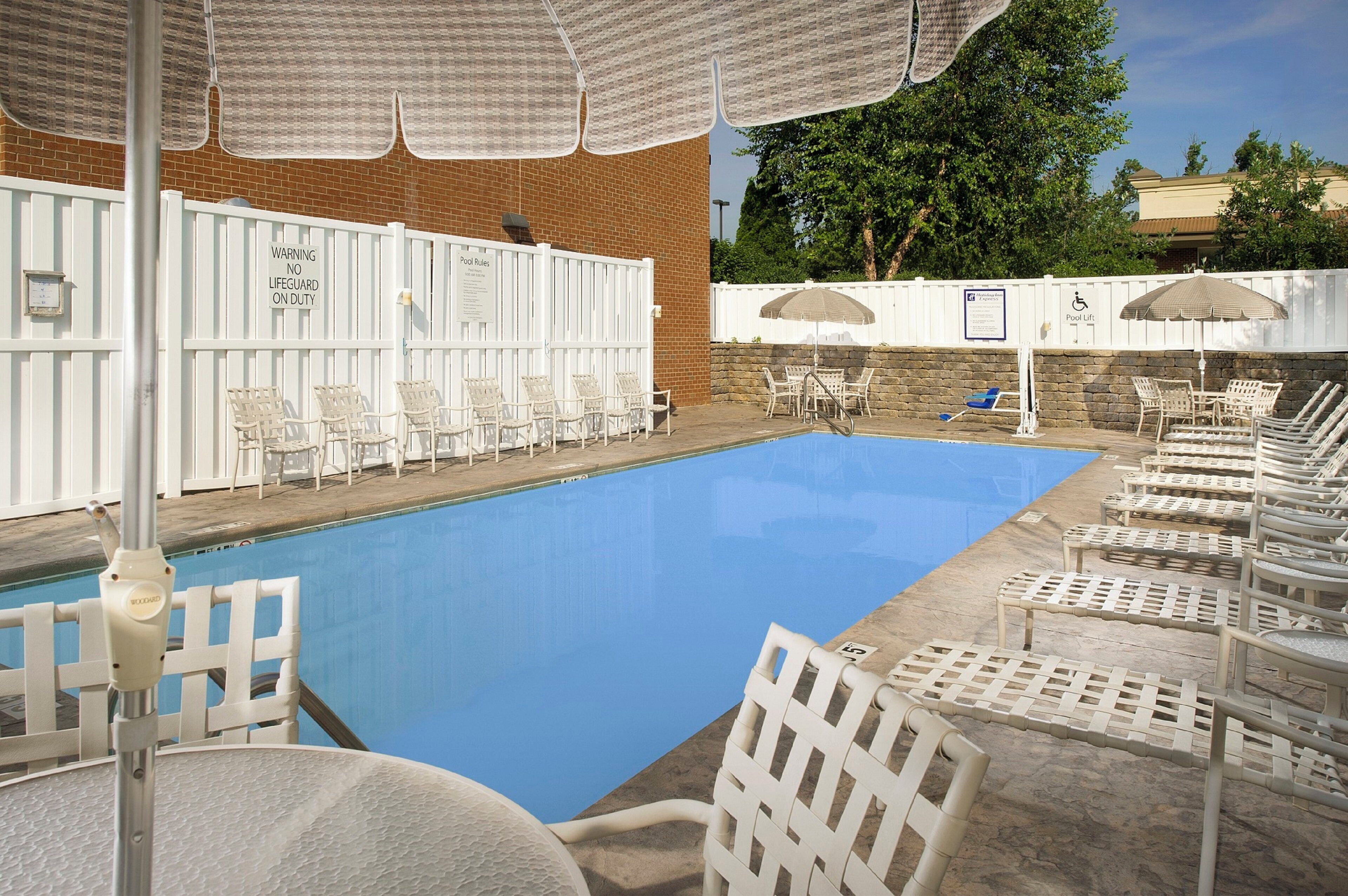 A swimming pool with lounge chairs and umbrellas, a white fence, and a brick wall.