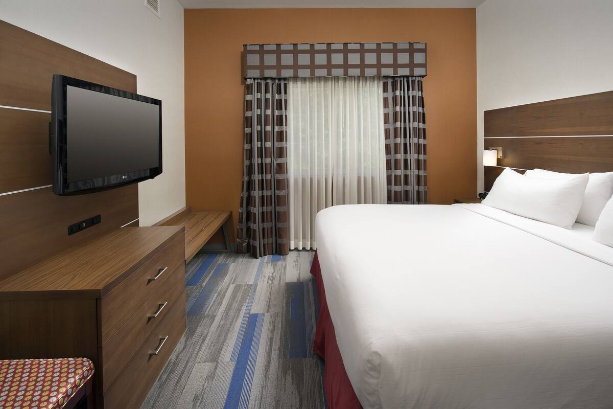A hotel room with a bed, a TV mounted on the wall, a desk with drawers, a bench, and a window with curtains.