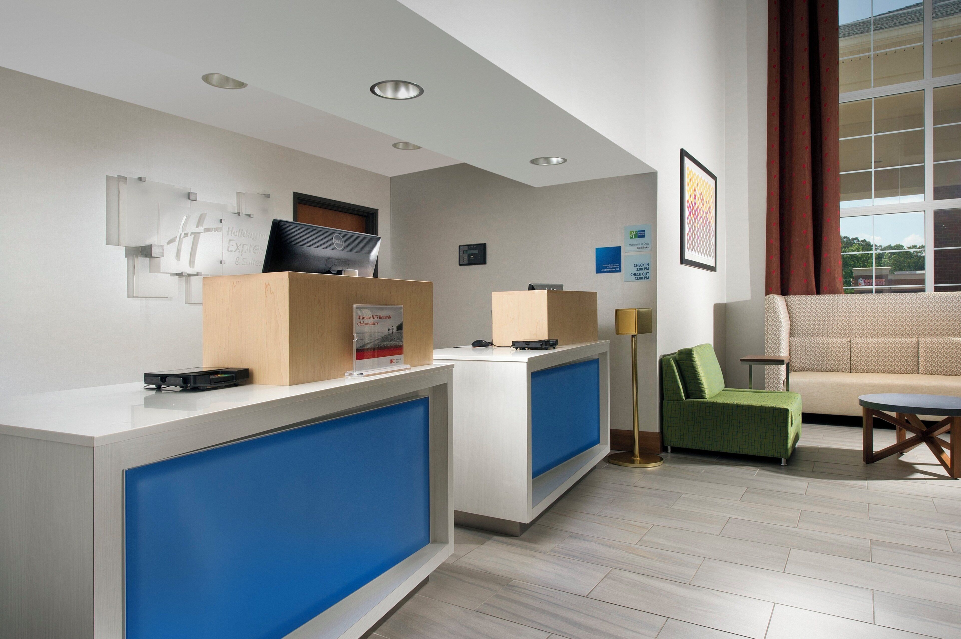 Zona de recepción de un hotel con mostrador, computadora y una mampara azul. Hay un sofá verde y una mesita con una lámpara.