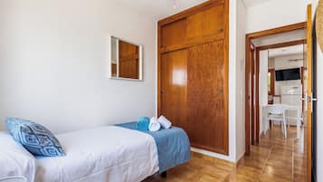 A bedroom with two beds, a wooden wardrobe, a mirror, and a view into the kitchen.
