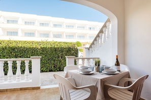 Un área de comedor al aire libre con sillas y mesas blancas, una botella de vino y vistas a un edificio blanco y un seto verde.